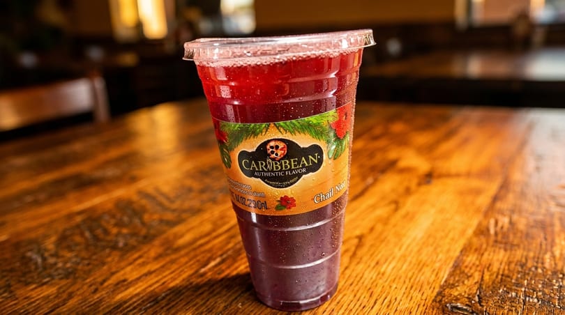 A Caribbean Authentic Flavor drink in a clear plastic cup with a lid on a wooden table.