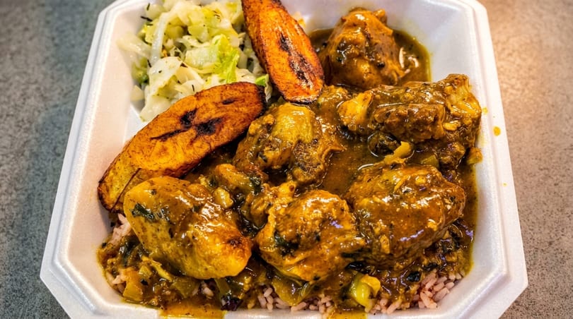 Curry chicken with rice, cabbage, and fried plantains in a white takeout container.