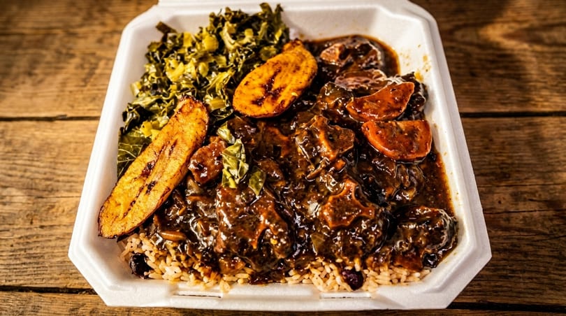 Oxtail stew with rice and peas, collard greens, and fried plantains in a takeout container.