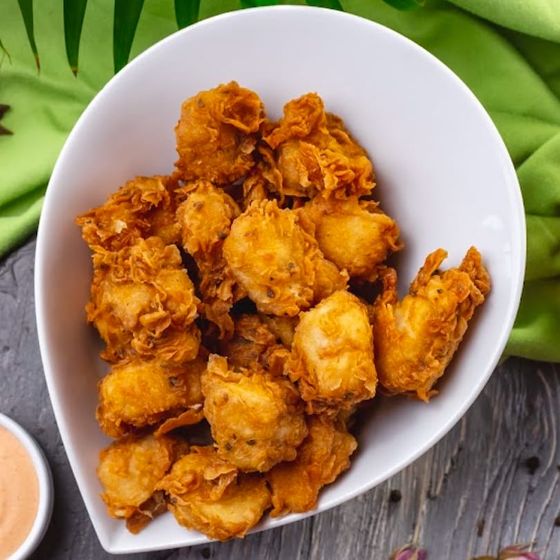 A white bowl filled with golden-brown Caribbean fried fritters, with a side of dipping sauce.
