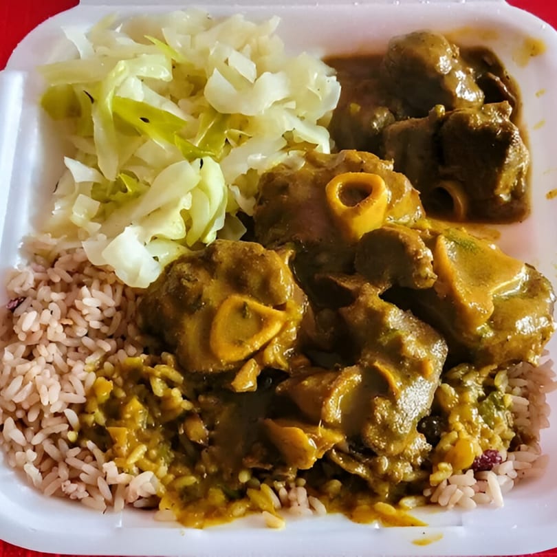 Curried goat with rice and peas, and steamed cabbage in a white takeout container.