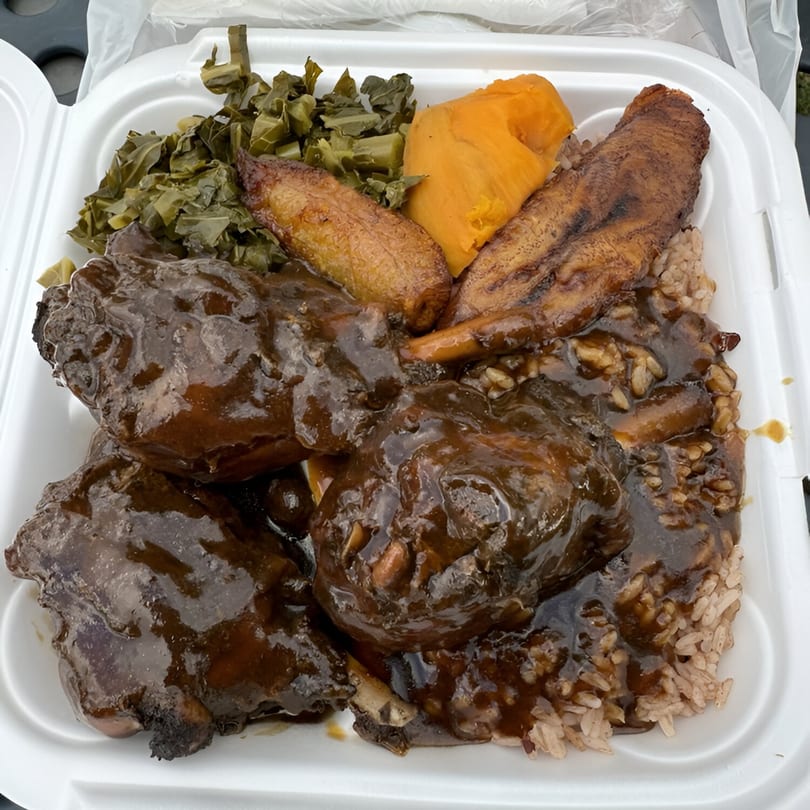 Caribbean oxtail with rice, collard greens, mashed pumpkin, and plantains in a takeout container.
